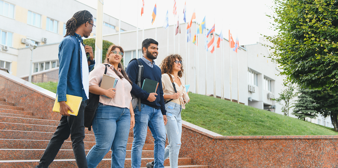 Four diverse students walking down outdoor steps carrying notebooks and backpacks near flags and a building.